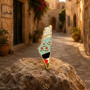 May include: A gold-toned metal enamel pin shaped like the map of Palestine. The design features a white keffiyeh pattern at the top, a light blue middle section with red flowers, and a bottom section with black, white, green, and red colors.