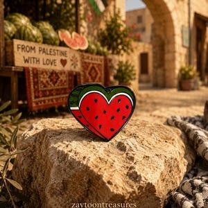May include: A heart-shaped enamel pin featuring a red watermelon slice with black seeds, a white rind, and a green outer edge. The design is rendered in vibrant colors with a glossy finish, showcasing a stylized fruit motif in a classic heart shape.