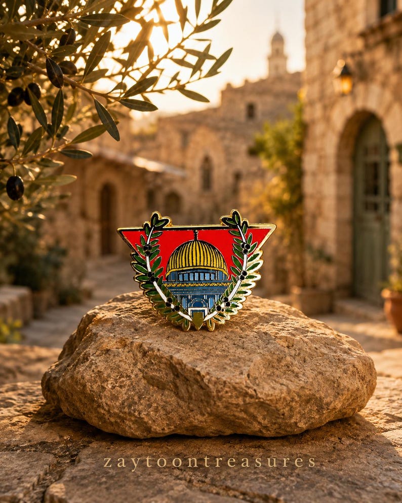 May include: A triangular enamel pin featuring a central illustration of the Dome of the Rock with a yellow dome and blue base, framed by two green olive branches on a vibrant red background, with gold-toned metal edges and a polished finish.