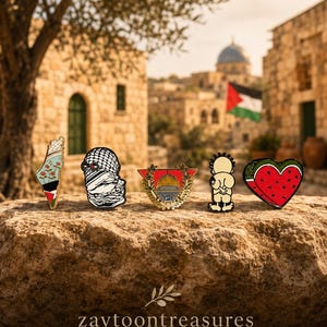 May include: Five enamel pins arranged in a row. The first is a map of Palestine with flowers and flag colors. The second is a keffiyeh-patterned figure. The third is a gold-framed Dome of the Rock. The fourth is a cartoon figure. The fifth is a watermelon heart.