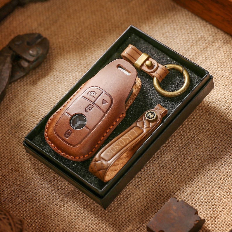 May include: A brown leather car key fob cover, key ring, and key holder set in a black box. The key fob cover has button icons and a keyhole. The key holder has the text "UK0808". The key ring is attached to a leather strap.