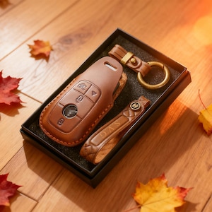 May include: A brown leather car key fob cover and matching key chain set in a black box. The key fob cover has button details and a key ring. The set is displayed on a wooden surface with autumn leaves.