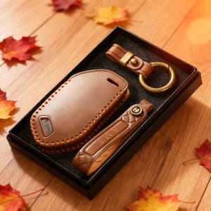 May include: A brown leather car key fob cover, key chain, and key fob holder set in a black box. The key fob cover is stitched and has a window. The key chain has a gold ring. Autumn leaves are scattered around the box.