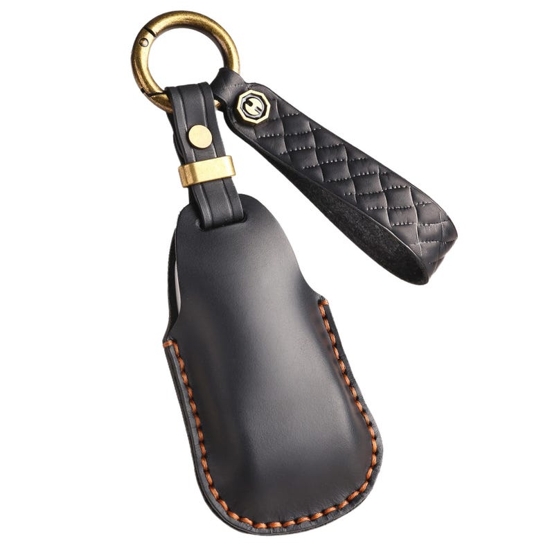 May include: Black leather key holder with a gold-colored ring and accents. The key holder has a stitched border and a quilted strap. The design includes a small, decorative emblem on the strap.