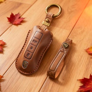 Puede incluir: Un juego de llavero y funda para llaves de cuero marrón con un anillo y herrajes dorados. La funda para llaves tiene botones en relieve y un logotipo de coche. El llavero tiene el número "116608" en relieve. Hojas de otoño dispersas.