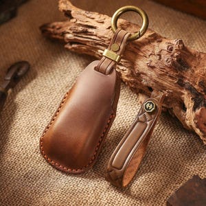 May include: Two brown leather keychains with brass hardware. One keychain is a rectangular case, and the other is a smaller strap with a decorative emblem. The keychains are on a textured surface with a piece of driftwood in the background.