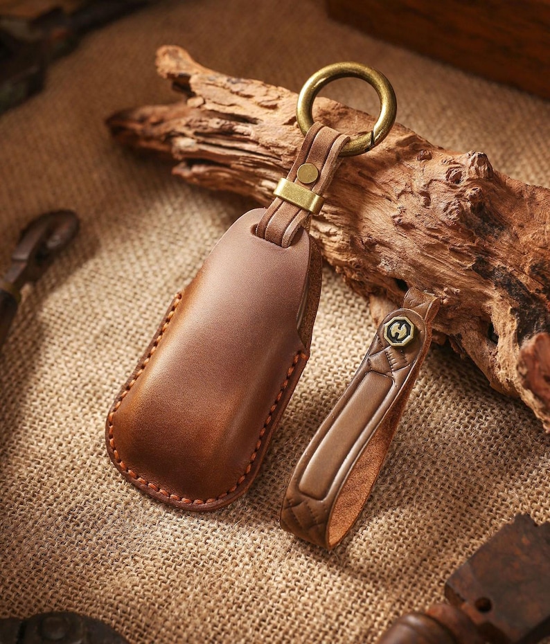 May include: Two brown leather keychains with brass hardware. One keychain is a rectangular case, and the other is a smaller strap with a decorative emblem. The keychains are on a textured surface with a piece of driftwood in the background.