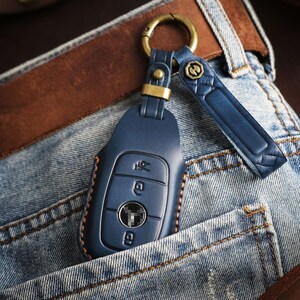 May include: A blue leather car key fob with a gold-colored ring and strap, resting in a denim pocket. The key fob has three buttons and is stitched with brown thread. A brown leather belt is visible in the background.