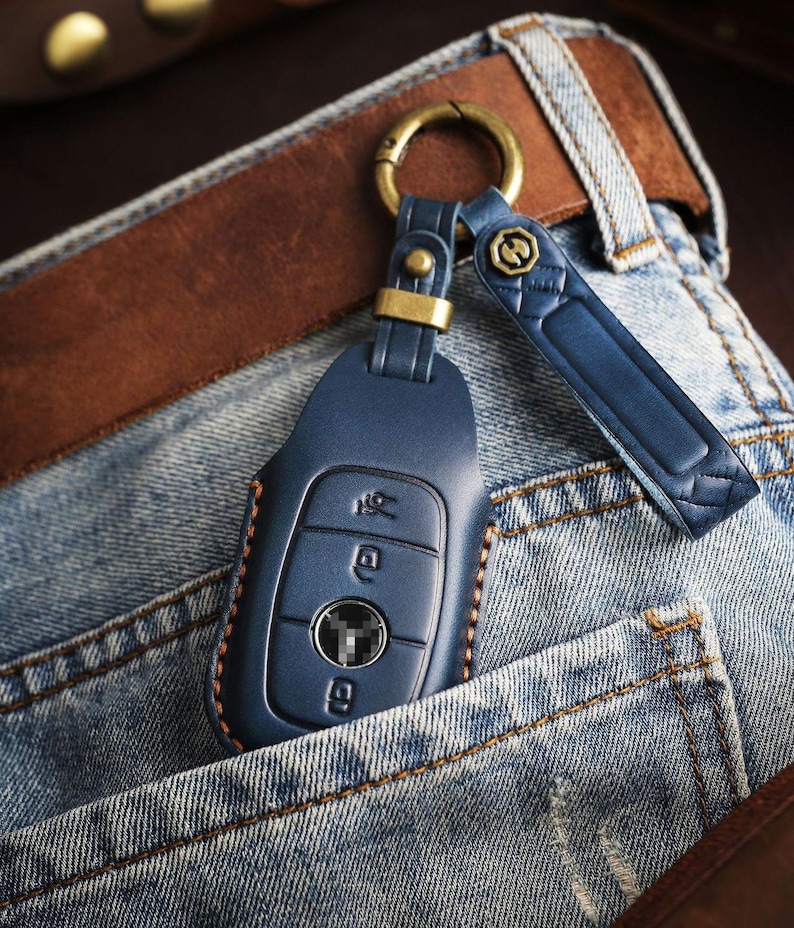 May include: A blue leather car key fob with a gold-colored ring and strap, resting in a denim pocket. The key fob has three buttons and is stitched with brown thread. A brown leather belt is visible in the background.