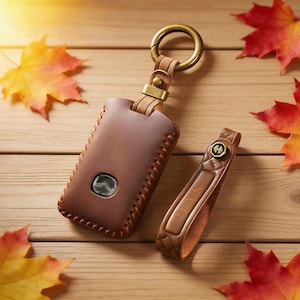 Pode incluir: Um porta-chaves e uma pulseira de couro castanho com um anel e ferragens douradas. O porta-chaves tem um design costurado e um emblema prateado. Folhas de outono vermelhas, laranja e amarelas estão espalhadas numa superfície de madeira.