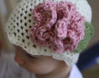 Peut inclure: Un bonnet bébé en crochet blanc avec une fleur en crochet rose et des feuilles vertes.