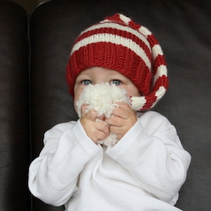 Puede incluir: Un niño con un gorro de punto rojo y blanco a rayas con una punta larga, sosteniendo un juguete de peluche blanco frente a su cara.