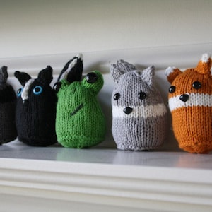 Op de afbeelding: Zes kleine gebreide dierenknuffels, waaronder een wasbeer, een stinkdier, een kikker, een eekhoorn, een vos en een kat. De knuffels zijn allemaal in verschillende kleuren en hebben zwarte ogen.