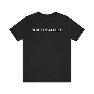 Może przedstawiać: Czarny t-shirt z białym napisem "SHIFT REALITIES" na piersi. Koszulka ma krótkie rękawy i okrągły dekolt. Tkanina wydaje się być miękkim, wygodnym materiałem.