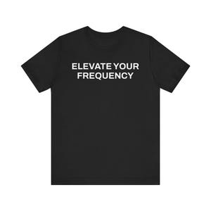 May include: Black t-shirt with the white text "ELEVATE YOUR FREQUENCY" across the chest. The short-sleeved shirt has a crew neck and is made of a soft material. The shirt is a unisex style.