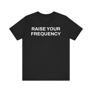 Puede incluir: Camiseta negra de cuello redondo con el texto blanco "RAISE YOUR FREQUENCY" en el pecho. La camiseta de manga corta está hecha de un material suave y es adecuada para ropa casual.
