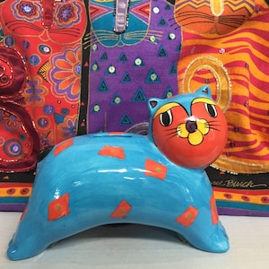 May include: A blue ceramic cat piggy bank with red and orange accents. The cat has a large, round head with a flower-shaped nose and large, black eyes. The cat is sitting on a colorful fabric background.