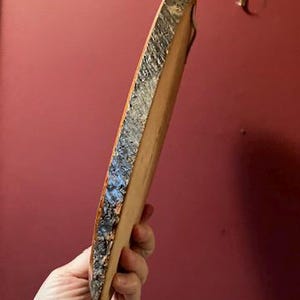 May include: A natural wood serving board with a live edge. The board is held at an angle, showcasing the rough, textured bark along one side and the smooth, light-colored wood on the other. A small, metal hook is visible in the background.