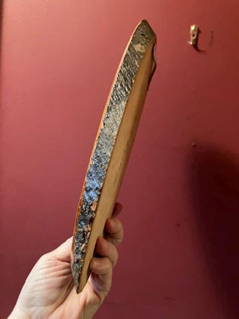 May include: A natural wood serving board with a live edge. The board is held at an angle, showcasing the rough, textured bark along one side and the smooth, light-colored wood on the other. A small, metal hook is visible in the background.