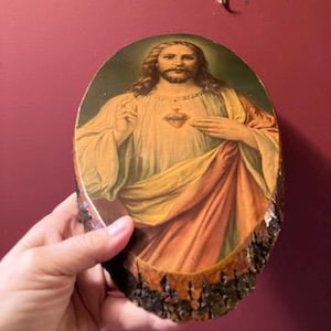 May include: Oval wooden plaque featuring a religious image of Jesus Christ. The artwork depicts Jesus with long hair, a beard, and a golden halo. He is wearing a cream-colored robe with orange and yellow draping. The edges of the plaque are the natural bark of the wood.