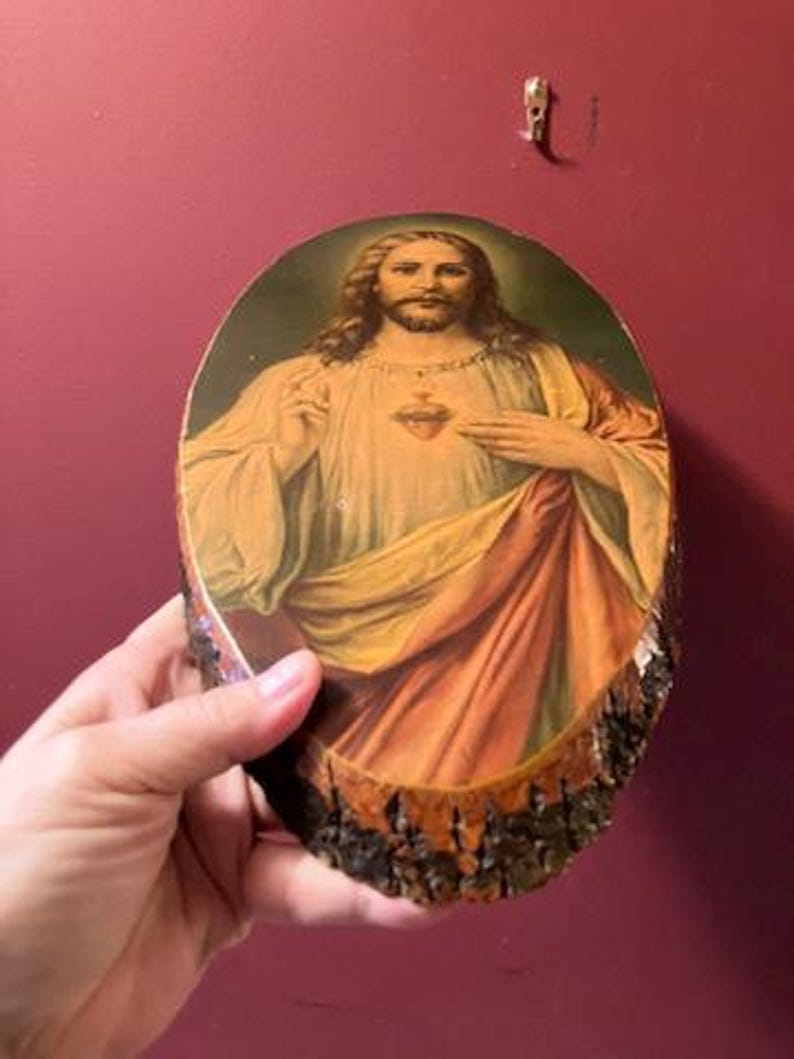 May include: Oval wooden plaque featuring a religious image of Jesus Christ. The artwork depicts Jesus with long hair, a beard, and a golden halo. He is wearing a cream-colored robe with orange and yellow draping. The edges of the plaque are the natural bark of the wood.
