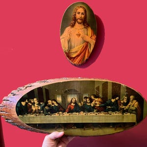 May include: Two religious artworks. The top piece is an oval-shaped portrait of Jesus with a golden halo and a red heart. The bottom piece is a long, irregular-shaped wooden plaque depicting The Last Supper.