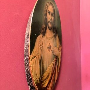 May include: Oval-shaped religious artwork depicting a figure with long hair and a beard, wearing a cream-colored robe and a red and gold drape. The artwork is framed with a rough, bark-like edge and is mounted on a pink wall.