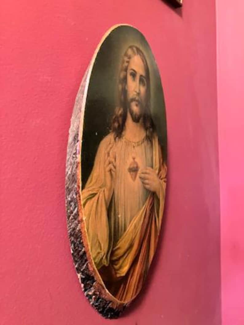May include: Oval-shaped religious artwork depicting a figure with long hair and a beard, wearing a cream-colored robe and a red and gold drape. The artwork is framed with a rough, bark-like edge and is mounted on a pink wall.
