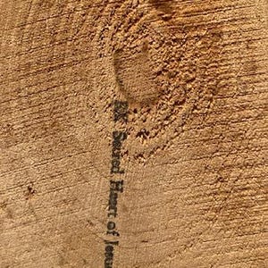 May include: Close-up of a section of wood with visible grain and a knot. The text "Sacred Heart of Jesus" is printed vertically in black ink on the wood's surface. The wood has a warm, natural brown color.