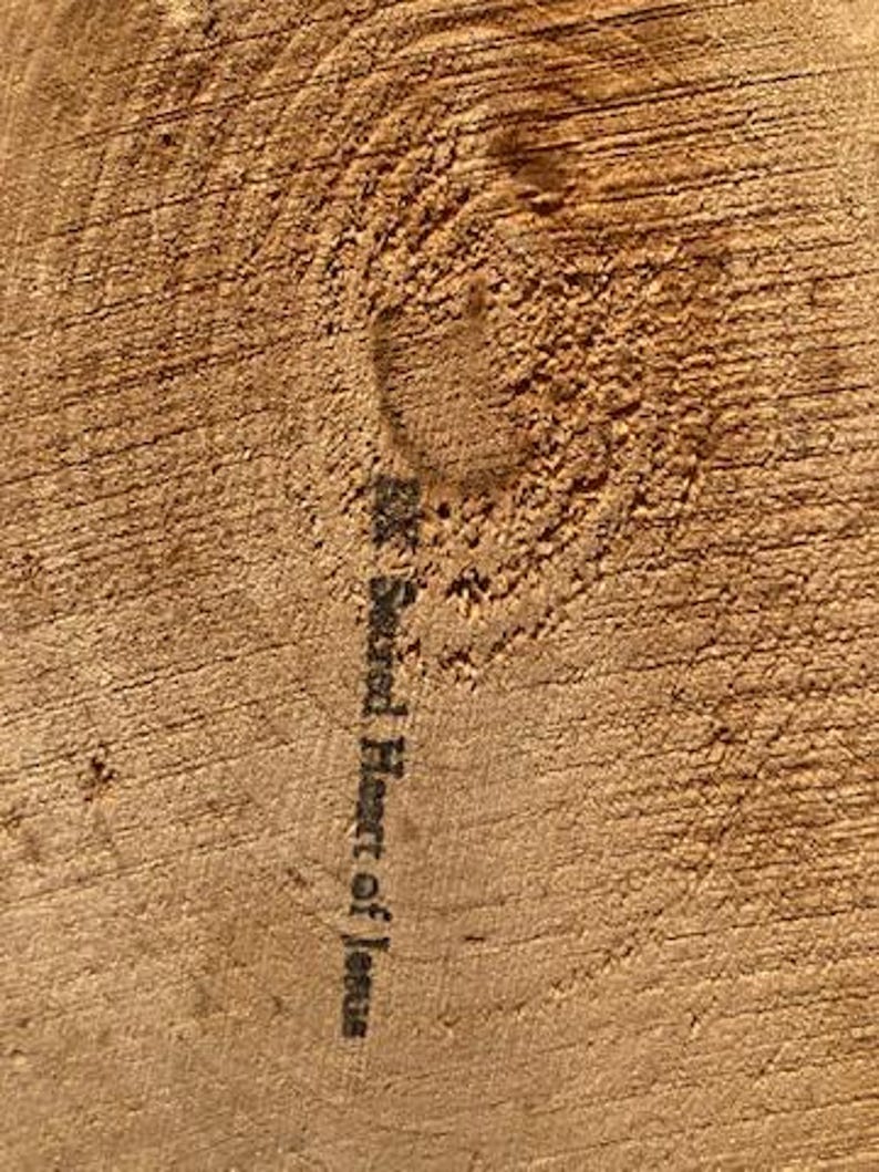 May include: Close-up of a section of wood with visible grain and a knot. The text "Sacred Heart of Jesus" is printed vertically in black ink on the wood's surface. The wood has a warm, natural brown color.