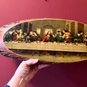 May include: An oval-shaped wooden plaque featuring a print of The Last Supper. The artwork depicts Jesus and his disciples seated at a long table. The wooden frame has a natural, rustic edge, and the background is a deep red.
