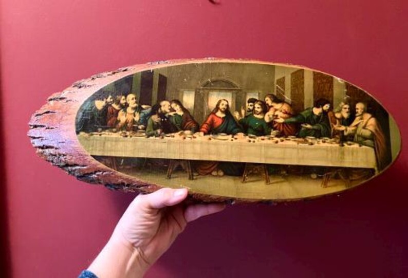 May include: An oval-shaped wooden plaque featuring a print of The Last Supper. The artwork depicts Jesus and his disciples seated at a long table. The wooden frame has a natural, rustic edge, and the background is a deep red.