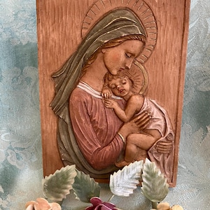 May include: A wooden relief sculpture of the Virgin Mary holding the baby Jesus. The sculpture is painted in shades of brown, pink, and green. The Virgin Mary is wearing a blue robe and a green veil. The baby Jesus is sleeping in her arms. The sculpture is framed in a wooden frame.