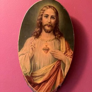 May include: Oval-shaped religious artwork depicting a figure with long brown hair and a beard, wearing a white robe and a red and yellow draped garment. A golden heart is visible on the chest. The artwork is framed by a natural wood edge.