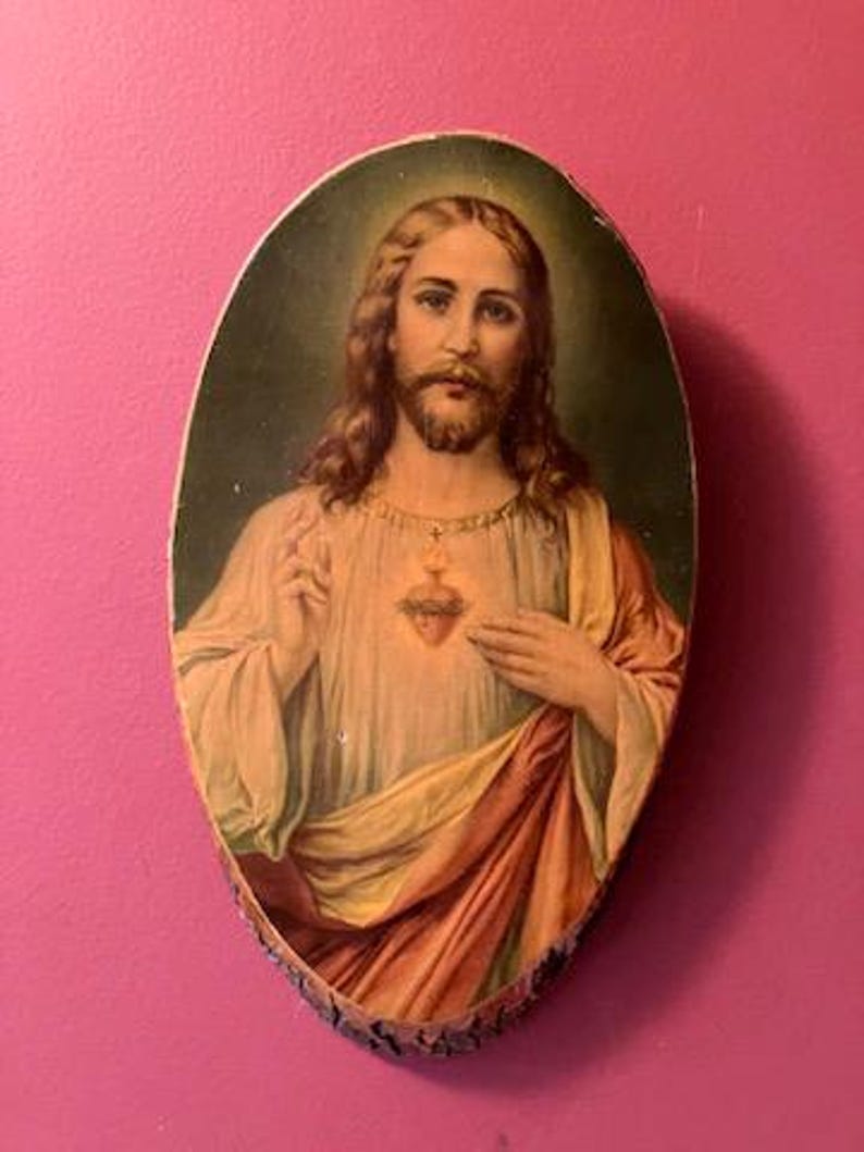 May include: Oval-shaped religious artwork depicting a figure with long brown hair and a beard, wearing a white robe and a red and yellow draped garment. A golden heart is visible on the chest. The artwork is framed by a natural wood edge.