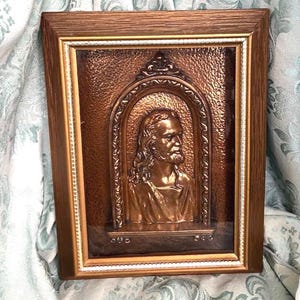 May include: A framed copper relief sculpture depicting a man with long hair and a beard. The artwork is set within a decorative arch and framed in a wooden frame with a gold inner border. The relief has a textured background.
