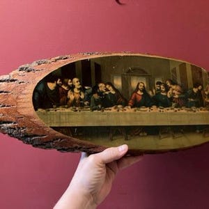 May include: An oval-shaped wooden plaque with a bark edge, featuring a print of The Last Supper. The artwork depicts Jesus and his disciples at a table, with a warm color palette. The plaque is held against a red background.