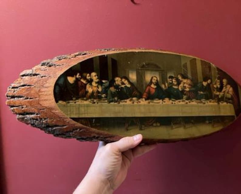 May include: An oval-shaped wooden plaque with a bark edge, featuring a print of The Last Supper. The artwork depicts Jesus and his disciples at a table, with a warm color palette. The plaque is held against a red background.