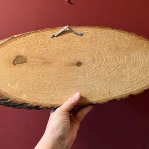 May include: Oval-shaped wooden plaque with a natural bark edge. The light brown wood grain is visible, and a metal hanger is attached to the top. The plaque is held by a hand, showcasing the wood's texture and color.