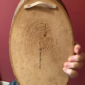 May include: A round, wooden wall hanging with a natural bark edge. The back features a beige fabric loop for hanging and a printed inscription. The wood grain is visible, and the piece is held by a person.
