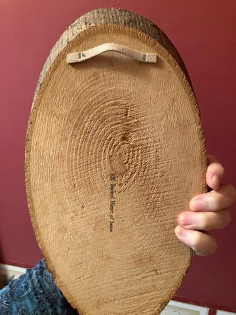 May include: A round, wooden wall hanging with a natural bark edge. The back features a beige fabric loop for hanging and a printed inscription. The wood grain is visible, and the piece is held by a person.