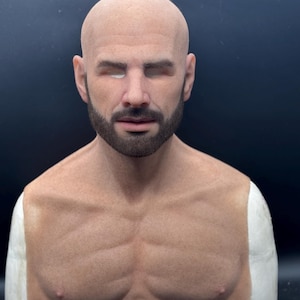 May include: A realistic, bald head mask with a short, dark beard and mustache. The mask features a detailed face with closed eyes and a sculpted chest. The skin tone is a light tan color, and the mask is set against a dark background.