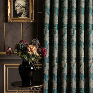 Könnte beinhalten: Elegante, teal- und goldfarbene Vorhänge mit Muster, ein gerahmtes Porträt und eine schwarze Vase mit Blumen. Die Vorhänge weisen ein detailliertes Paisley-Design auf und verleihen der Raumdekoration einen Hauch von Luxus. Ein goldener Beistelltisch mit schwarzer Platte ist ebenfalls zu sehen.
