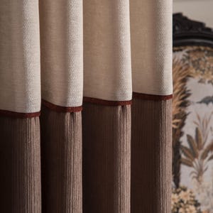 Può includere: Primo piano di eleganti tende a strati. La sezione superiore è in tessuto beige chiaro, che sfuma in un velluto a coste marrone scuro nella parte inferiore. Un sottile bordo rosso scuro separa le due sezioni. Lo sfondo mostra una sedia fantasia.