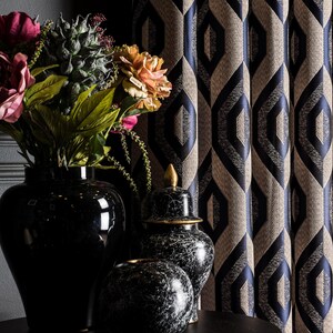 May include: A still-life arrangement featuring a black vase with colorful flowers, a decorative black and white marble jar, and a patterned curtain with a geometric design in navy blue and beige. The scene is set against a dark gray wall.