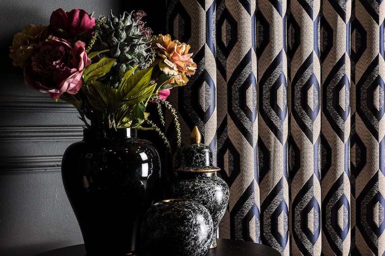 May include: A still-life arrangement featuring a black vase with colorful flowers, a decorative black and white marble jar, and a patterned curtain with a geometric design in navy blue and beige. The scene is set against a dark gray wall.