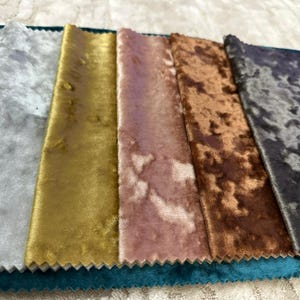 K&ouml;nnte beinhalten: Eine Sammlung rechteckiger Stoffmuster mit einer Knitter-Samt-Textur. Die Farben umfassen Hellgrau, Gold, Altrosa, Kupfer und ein dunkles, meliertes Grau, die nebeneinander auf einem t&uuml;rkisfarbenen Grundstoff mit gezackten Kanten angeordnet sind.