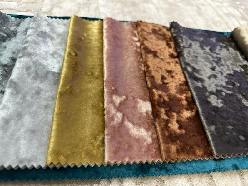K&ouml;nnte beinhalten: Eine Sammlung rechteckiger Stoffmuster mit einer Knitter-Samt-Textur. Die Farben umfassen Hellgrau, Gold, Altrosa, Kupfer und ein dunkles, meliertes Grau, die nebeneinander auf einem t&uuml;rkisfarbenen Grundstoff mit gezackten Kanten angeordnet sind.