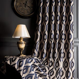 May include: A room interior featuring a patterned curtain, table cloth, and a wall clock. The curtain and table cloth have a repeating geometric design in navy blue and beige. A gold-toned lamp sits on the table. A round clock hangs on the wall.