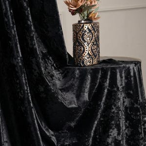 K&ouml;nnte beinhalten: Ein schwarzer Vorhang aus Knitter-Samt mit tiefem, strukturiertem Flor. Neben dem Stoff steht eine zylindrische Vase mit einem metallisch goldenen und schwarzen Ornamentmuster, die einen Strau&szlig; aus wei&szlig;en und pfirsichfarbenen Kunstblumen enth&auml;lt.